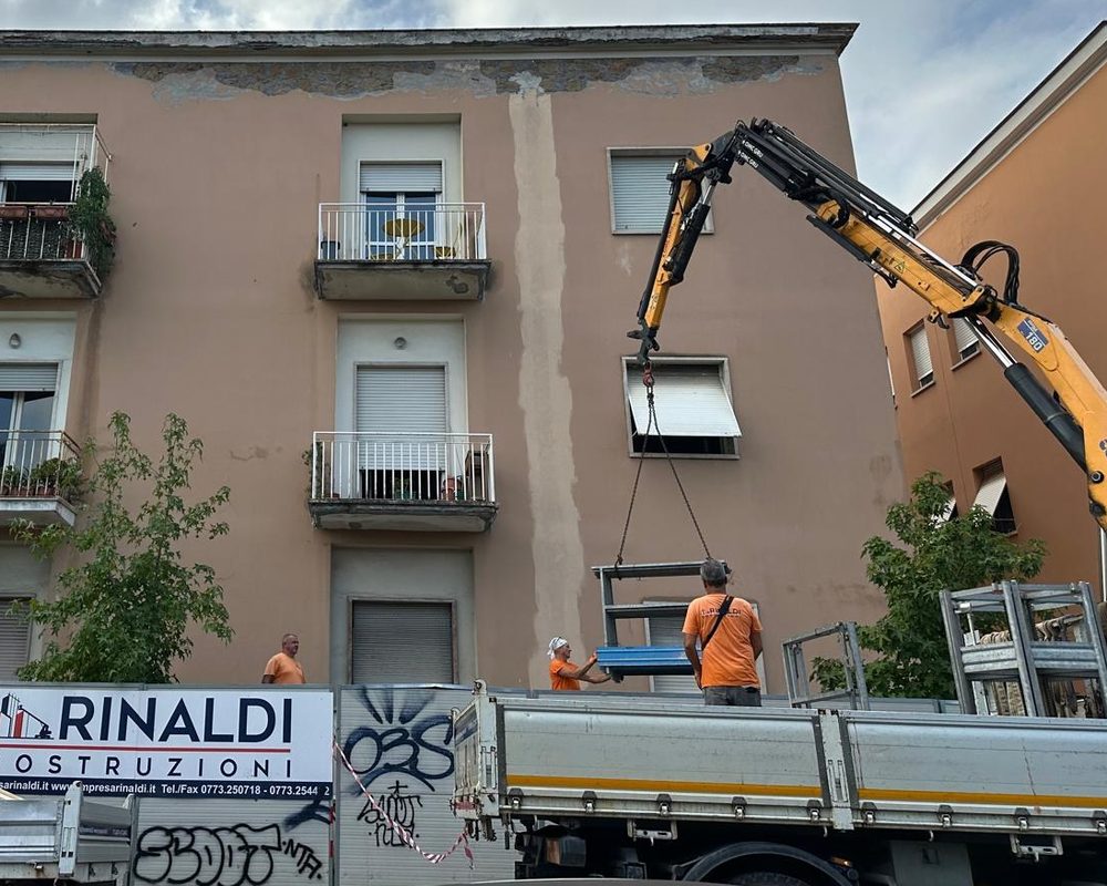 Gru su autocarro Copma che solleva materiali davanti a un condominio in via Don Minzoni, con gli operai Rinaldi