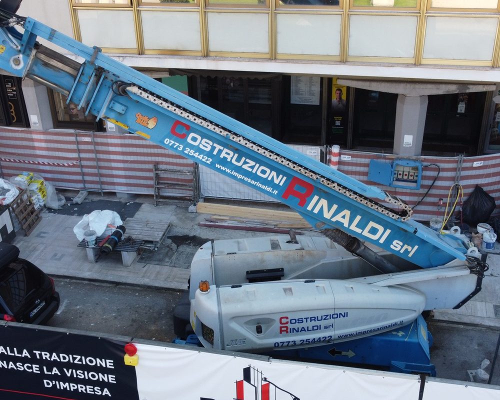 Piattaforma aerea JLG con la scritta Costruzioni Rinaldi sul braccio, vista dal drone in un cantiere a Latina